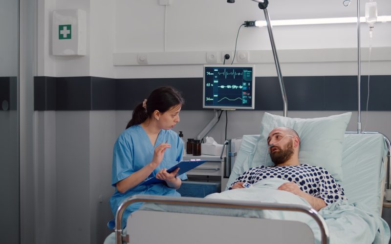 Medical nurse discussing illness treatment with hospitalized sick man resting in bed during rehabilitation therapy in hospital ward. Patient with nasal oxygen tube explaining disease symptom